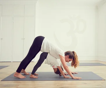 "Adult and child practicing yoga in downward dog pose on mats in a bright room with an Om symbol on the wall."