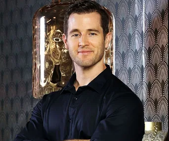 Man in black shirt smiling with arms crossed in front of patterned wallpaper and unique lamp in background.