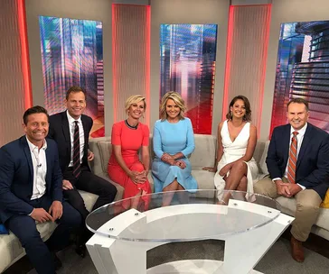 Group of six TV hosts sitting on a morning show set, featuring bright colorful attire and a glass coffee table in front.