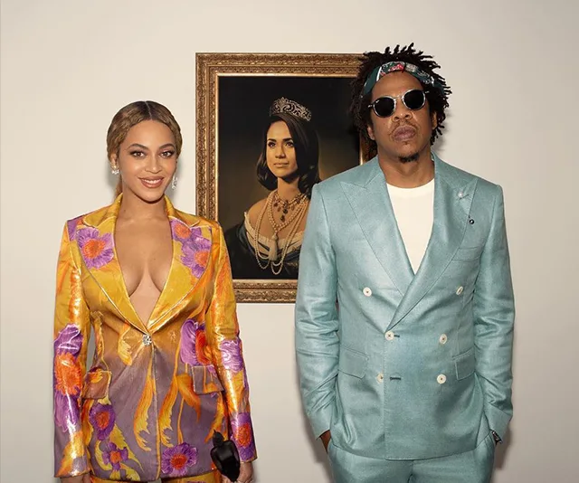Two individuals in colorful suits stand beside a portrait of a woman in a tiara and necklace.