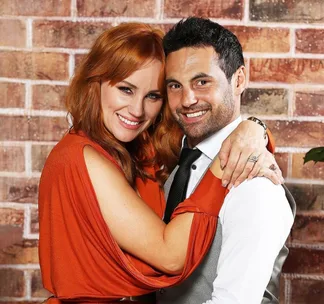Couple embracing and smiling in front of a brick wall. Woman in orange dress, man in dress shirt and tie.
