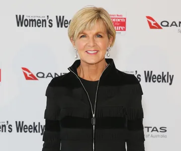 A woman in a black jacket smiles on the red carpet at a Women's Weekly event with logos in the background.