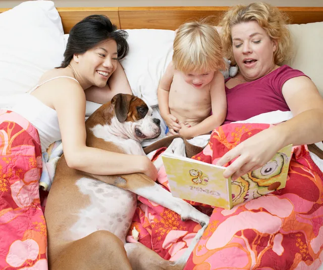 A family, including two women, a child, and a dog, reading a book together in bed under a colorful blanket.