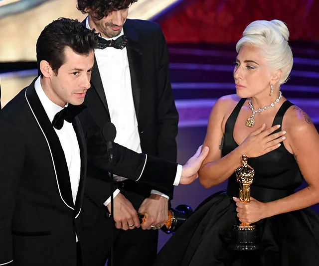 Oscar winners on stage, holding awards, lady in black dress with blonde hair, man in black tuxedo giving speech.