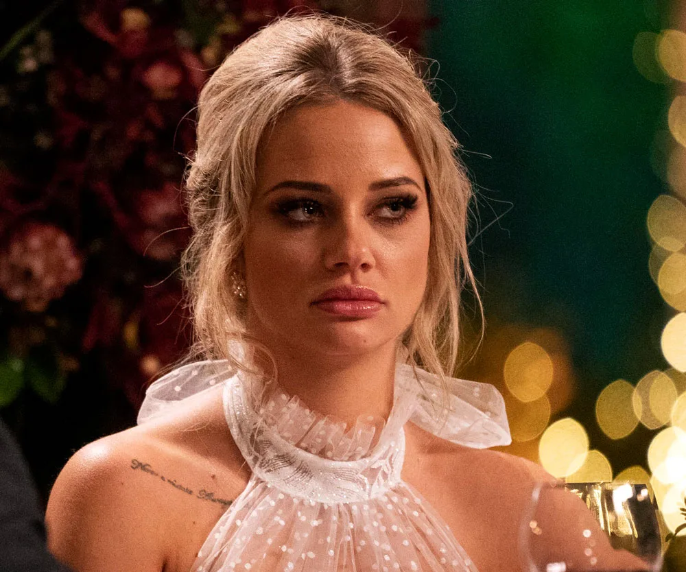 A woman with blonde hair in a white outfit looking serious, in front of a blurred festive background.