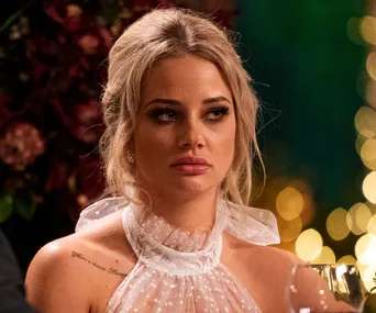 A woman with blonde hair in a white outfit looking serious, in front of a blurred festive background.
