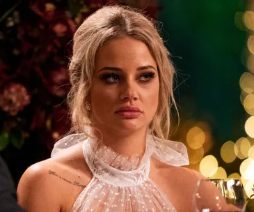 A woman with blonde hair in a white outfit looking serious, in front of a blurred festive background.