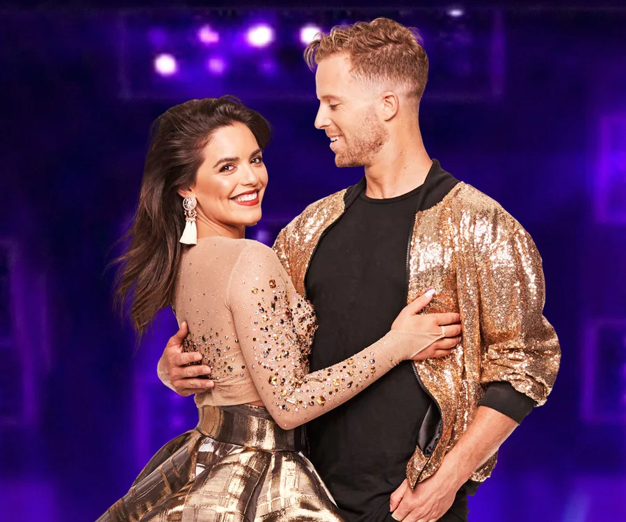 Couple in sparkling outfits smiling against a purple backdrop, possibly from a dance show.