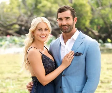 A couple dressed formally poses outdoors; the woman in a dark dress, the man in a light blue suit, both smiling.