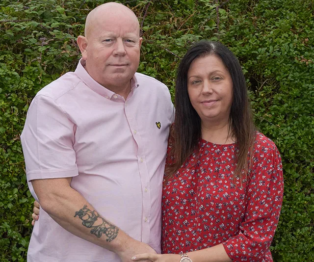 A man in a pink shirt with a tattoo and a woman in a red floral dress stand together in front of greenery.