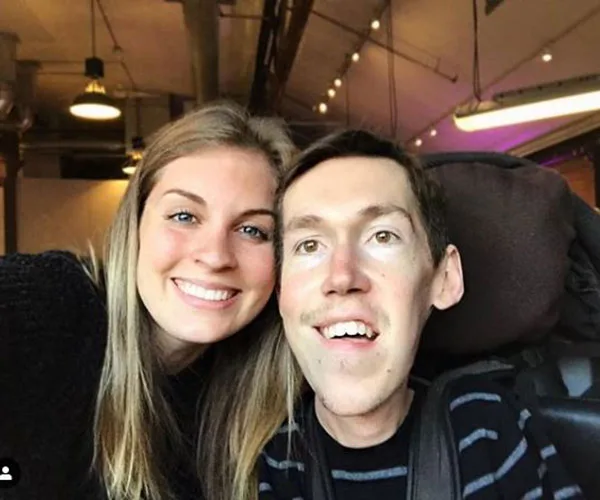 A smiling couple, with the man in a wheelchair, pose together in a warmly lit indoor setting.