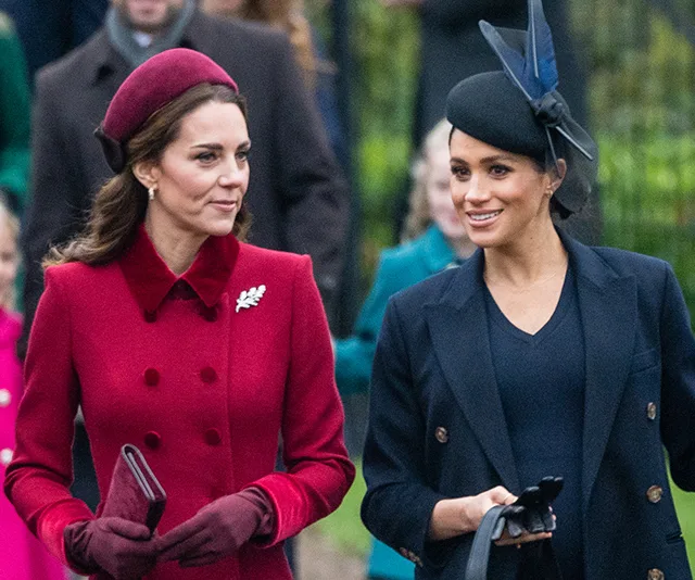 Two women walking outdoors, one in a red coat and beret, the other in a black coat with a hat, both smiling.