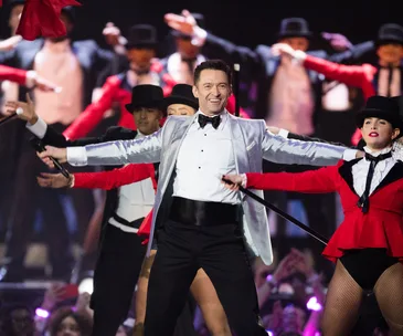 Hugh Jackman performs energetically on stage in a silver tuxedo, surrounded by dancers in red and black costumes.