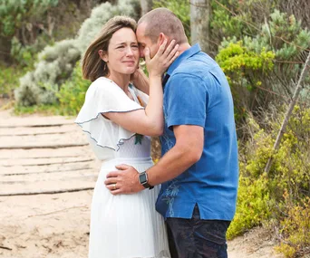A man comforts a woman as she cries on a sandy pathway surrounded by greenery.
