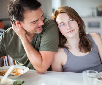 A father and teenage daughter share a light-hearted moment at the dining table, with a partially eaten meal in view.