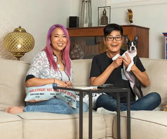 Couple seated on couch, woman with purple hair holding a menu, man holding a Boston Terrier, living room setting.