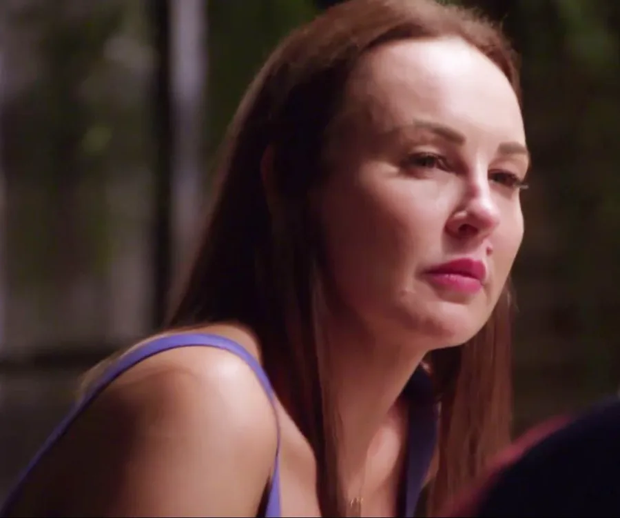 A person with long brown hair and a serious expression, wearing a blue tank top, in a dimly lit setting.