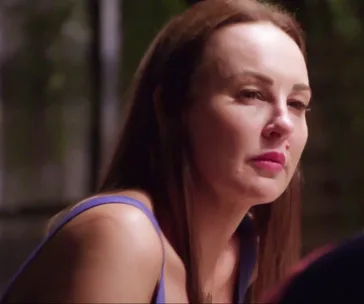 A person with long brown hair and a serious expression, wearing a blue tank top, in a dimly lit setting.