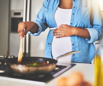 Pregnant woman in a blue shirt cooking with a pan on a stove, holding a wooden spoon in a kitchen setting.