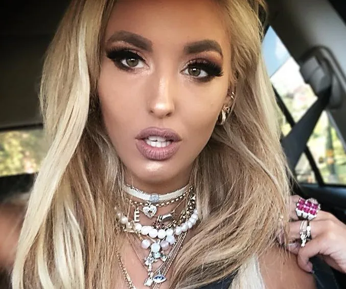 Alt text: "Woman with long blonde hair wearing layered necklaces and pink ring, seated in a car."