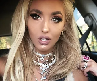 Alt text: "Woman with long blonde hair wearing layered necklaces and pink ring, seated in a car."