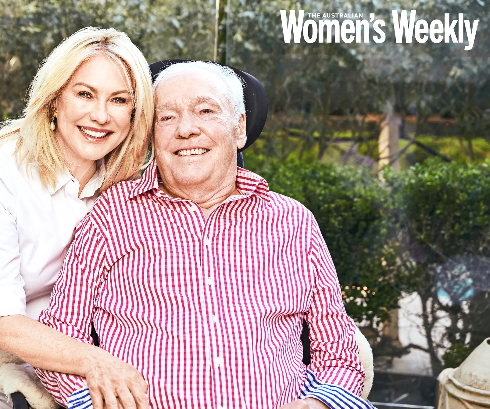 A smiling woman poses with an older man in a wheelchair, outdoors with greenery in the background.