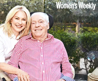A smiling woman poses with an older man in a wheelchair, outdoors with greenery in the background.