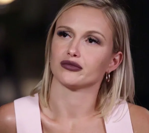 Young woman with blonde hair and hoop earrings, wearing a pink top, appears deep in thought.