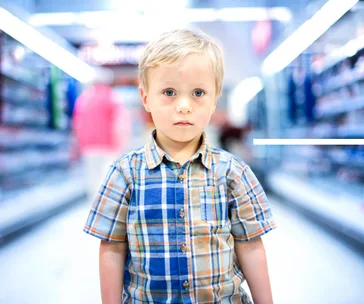 Child in a plaid shirt stands in a store aisle, looking directly at the camera, with shelves blurred in the background.