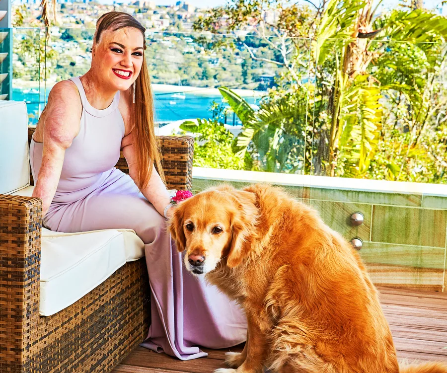 Woman in a lilac dress sitting outdoors with a golden retriever by her side, lush greenery in the background.