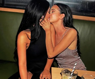 Two women seated on a bench embrace and kiss in a dimly lit bar setting.