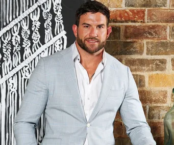 Man in light blue suit with white shirt, standing against a brick wall backdrop.