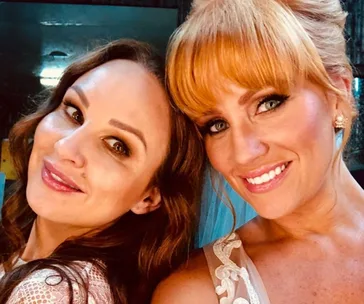 Two women smiling closely together, both with makeup and styled hair, in a warmly lit setting.