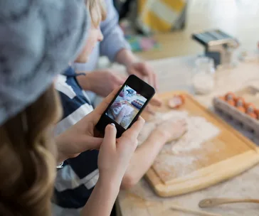 A child is guided in making dough on a board while another person photographs the process with a smartphone.
