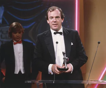 A man in a tuxedo holds an award on stage, with another man in a suit and red bow tie standing behind him.