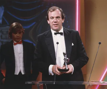 A man in a tuxedo holds an award on stage, with another man in a suit and red bow tie standing behind him.