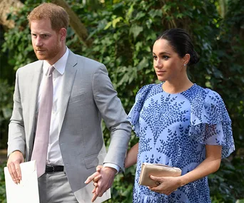 Prince Harry and Meghan Markle walking hand in hand outdoors, surrounded by greenery.