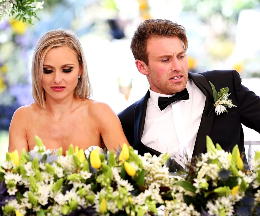 Man and woman in formal attire sitting at a table decorated with floral arrangements, both looking downwards.