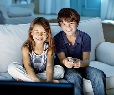 Two children smiling and sitting on a white couch, watching TV together.