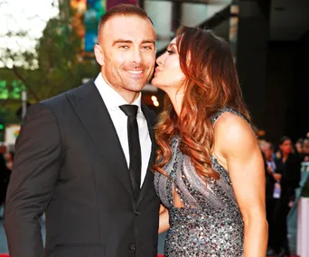 A couple on a red carpet, a woman in a sparkling dress kisses a smiling man in a suit.