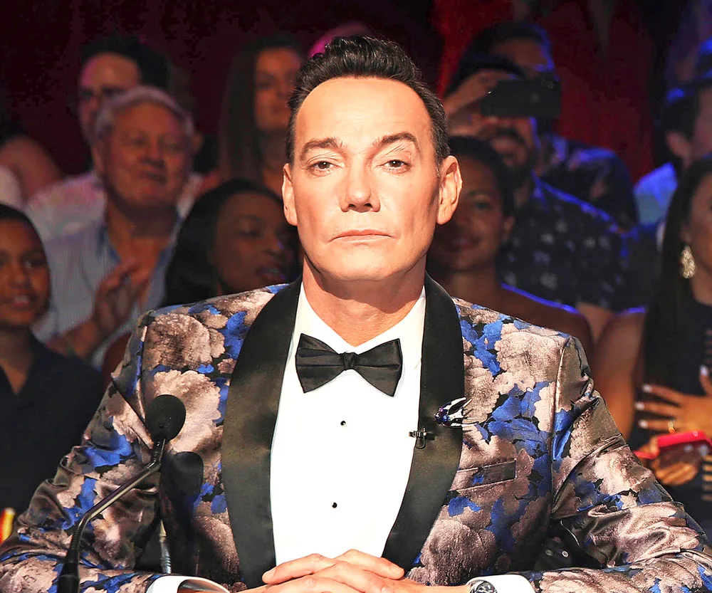 Judge in a floral blazer and bow tie sits at a table with a focused expression, audience blurred in the background.