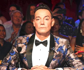 Judge in a floral blazer and bow tie sits at a table with a focused expression, audience blurred in the background.