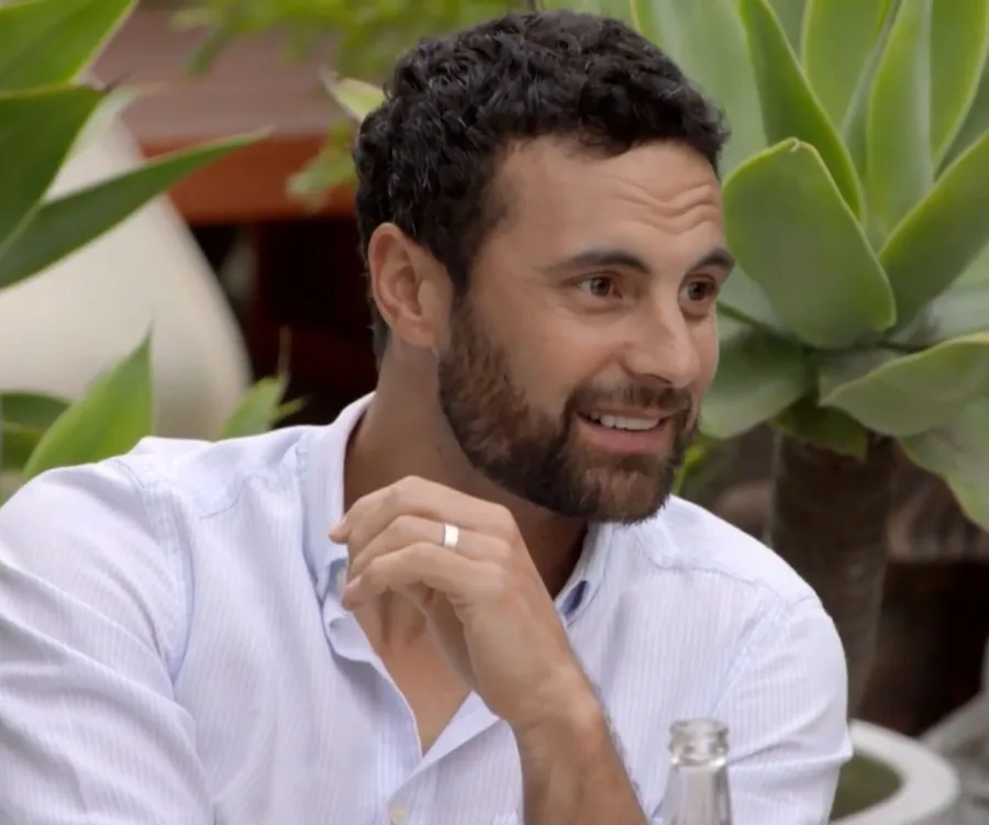 Man with beard in a white shirt, smiling in a garden setting with plants in the background.