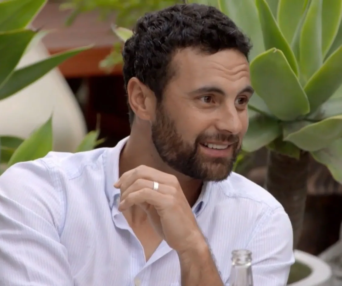 Man with beard in a white shirt, smiling in a garden setting with plants in the background.