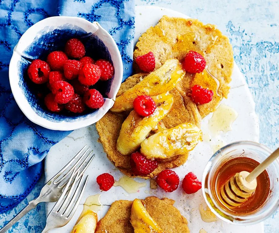 Wholemeal pancakes topped with caramelized banana, fresh raspberries, and honey on a blue-textured background.