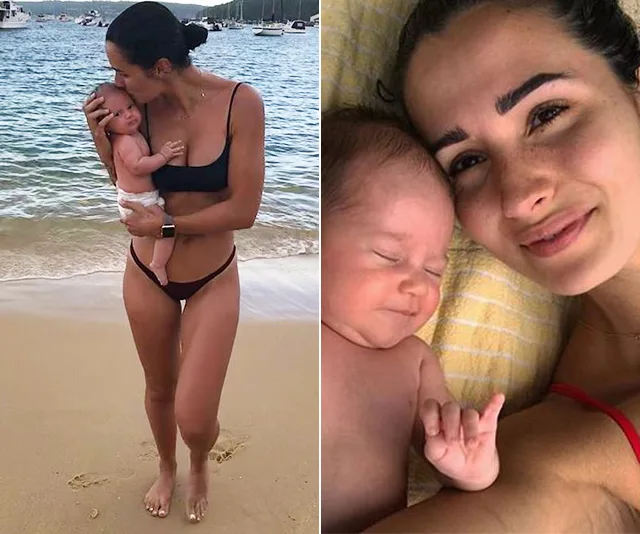 A woman in a bikini holds a baby on the beach and smiles with the baby indoors.