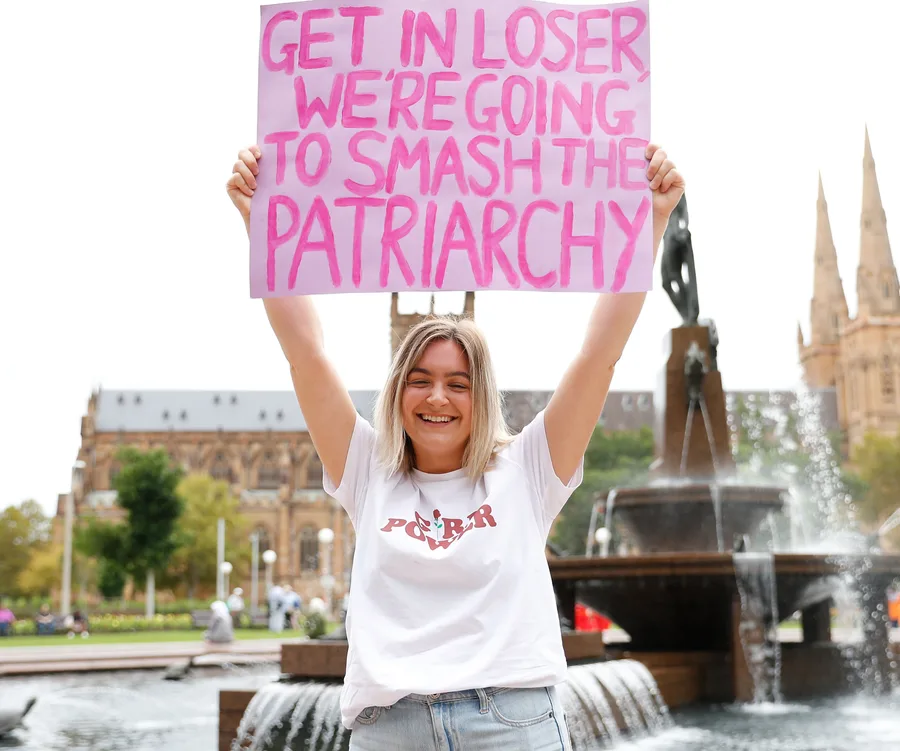 Woman smiling, holds pink sign saying "Get in loser, we're going to smash the patriarchy" in front of a cathedral.
