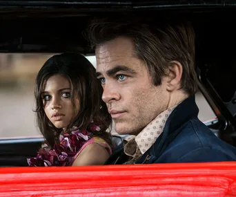 Two individuals sitting in a car, one wearing a floral lei, both with serious expressions.