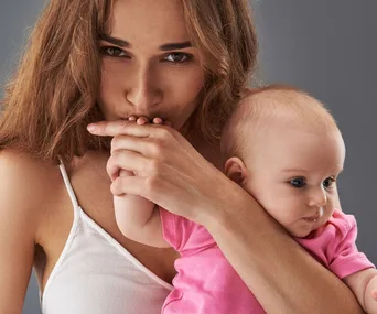 Woman kisses a baby's hand while holding the baby, both wearing casual clothing against a gray background.