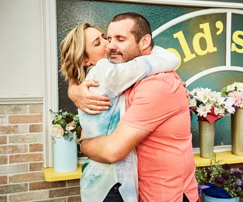 A man and woman embrace affectionately outside a flower shop with yellow decor and various floral arrangements.